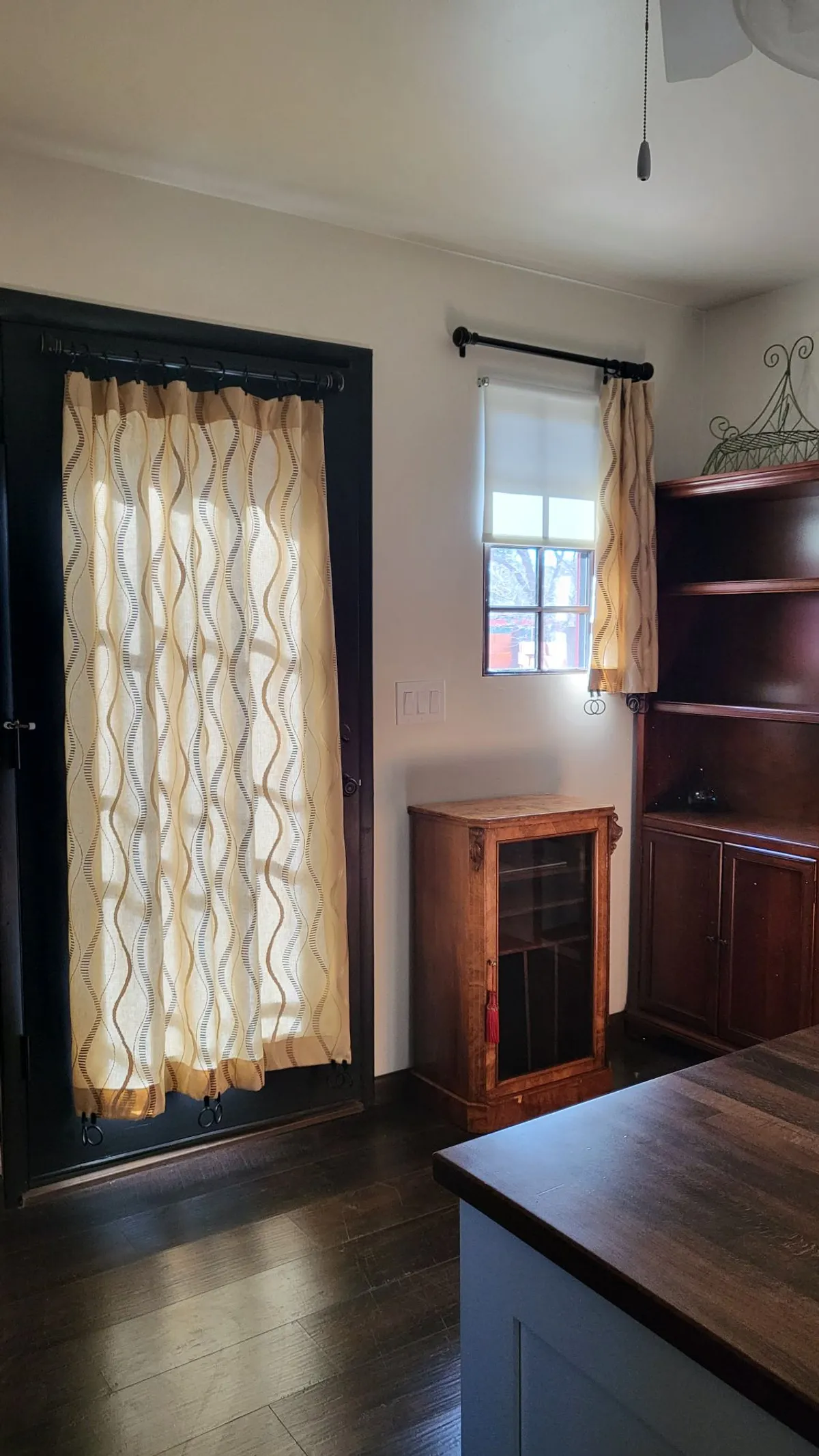 A cozy nook with a wooden cabinet, bookshelf, and soft curtains. Sunlight streams through the window, illuminating the reading and relaxing area. The arrangement suggests a calm, private retreat ideal for curling up with a book or enjoying a peaceful moment at home.