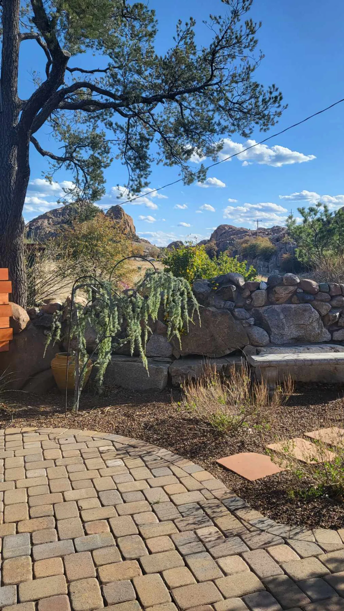 This image captures a peaceful outdoor scene featuring a large tree, a dramatic rock wall, a paved path, a comfortable bench, and a sweeping landscape view. The setting suggests a private and tranquil area ideal for relaxing, meditating, or simply enjoying the surrounding natural beauty. The elements visible convey a seamless transition from the comfort of home to the adventure and serenity of the outdoors.